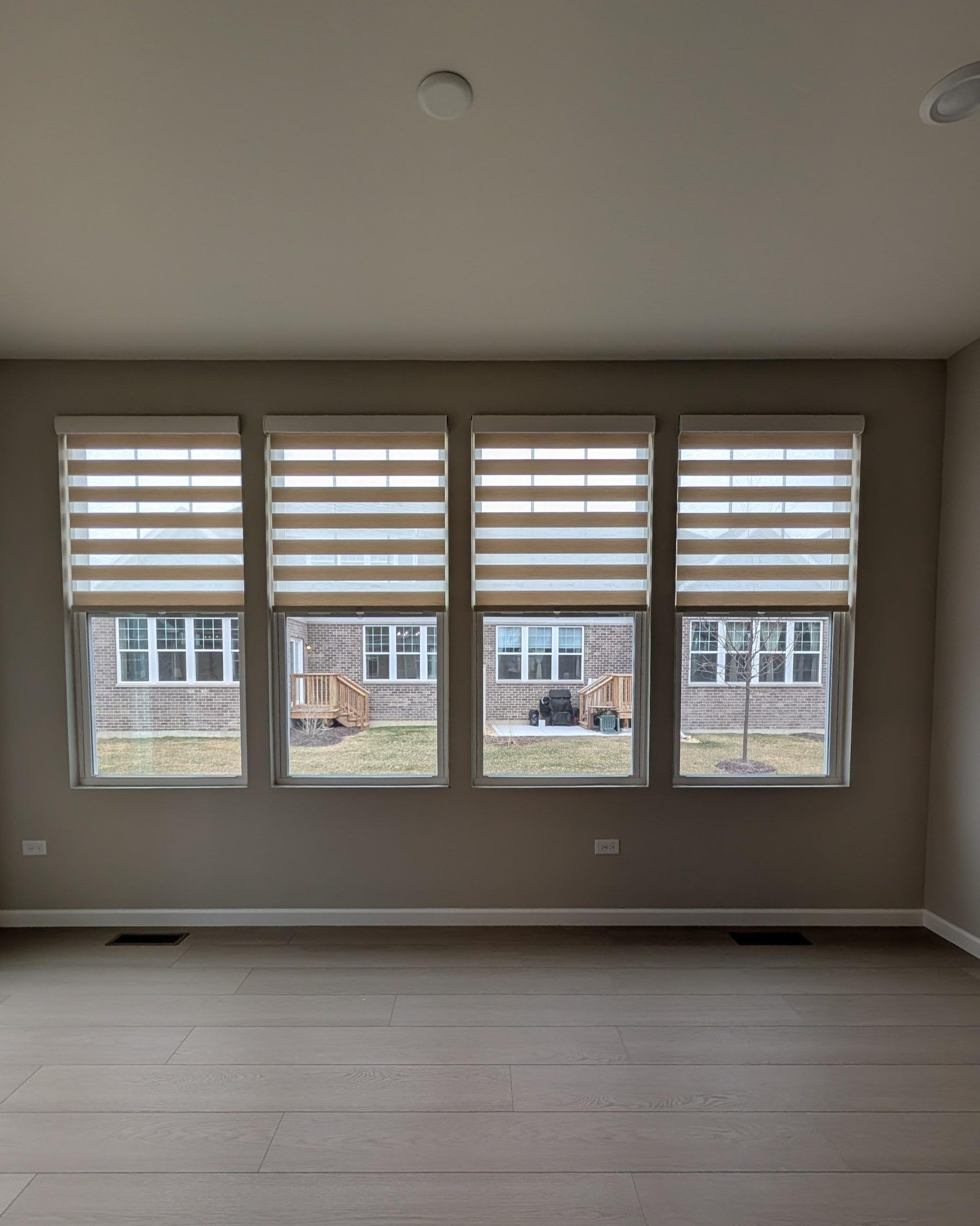 Empty living room with four tall windows covered by horizontal blinds, beige walls, and light wood flooring. Outside, brick houses and a small yard are visible.