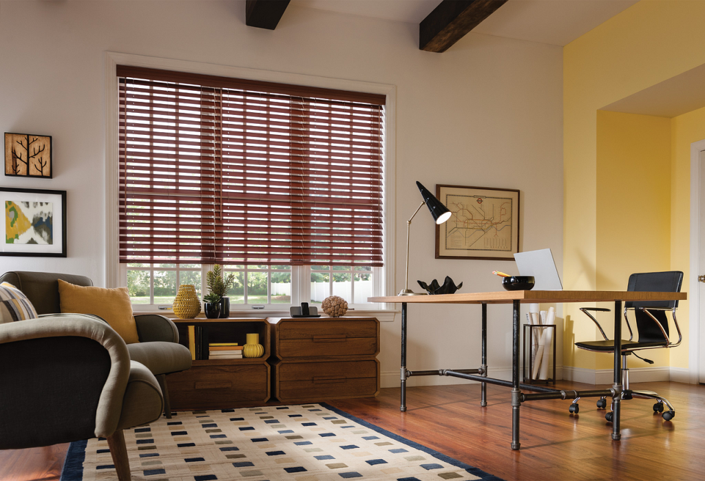 Sunlit home office with a wooden desk, laptop, and black task lamp near a window with brown blinds.
