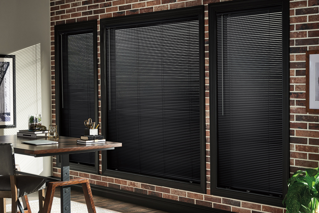 Home office with brick wall and three tall windows covered by black horizontal blinds; a wooden desk and chair sit to the left.